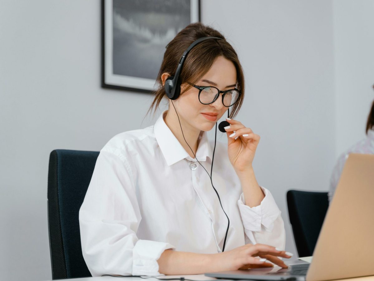 Young woman in glasses and headset providing customer support at a laptop in an office setting.